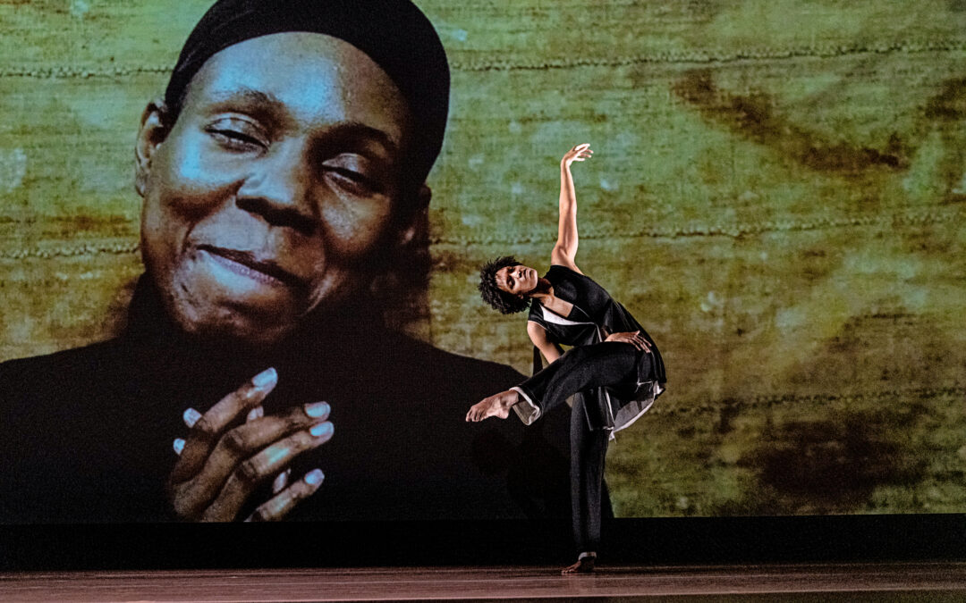 a female dancer wearing black performing in front a large projecting of a Black man smiling