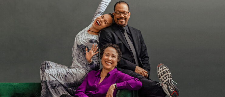 Tai Jimenez, Robert Garland, and Virginia Johnson pose together seated on a green velvet couch. Jimenez, perched on the back, leans back against Garland as she stretches an arm overhead, posing as though she's singing karaoke. Garland wears a wry smile as he kicks a sneakered foot out towards the camera. Seated below them, Johnson smiles warmly, exuding elegance and calm.