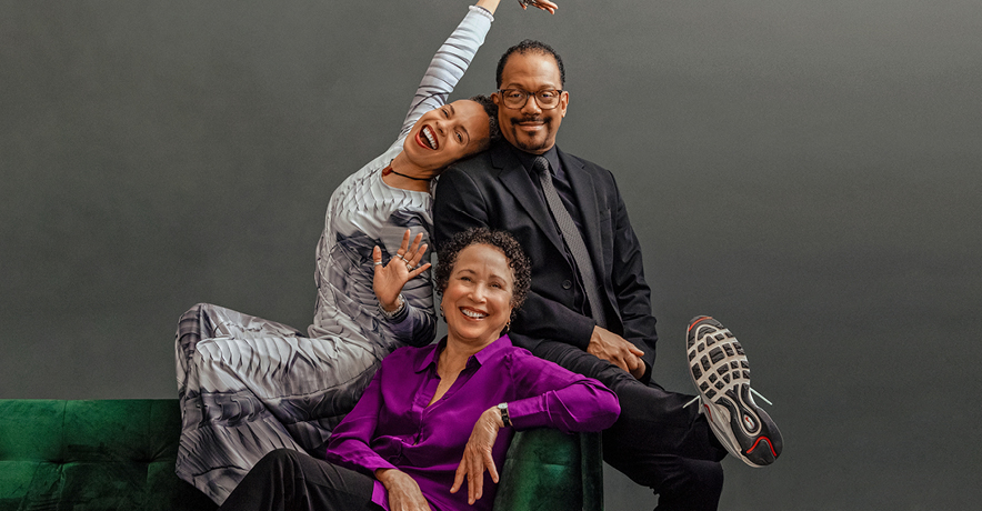 Tai Jimenez, Robert Garland, and Virginia Johnson pose together seated on a green velvet couch. Jimenez, perched on the back, leans back against Garland as she stretches an arm overhead, posing as though she's singing karaoke. Garland wears a wry smile as he kicks a sneakered foot out towards the camera. Seated below them, Johnson smiles warmly, exuding elegance and calm.