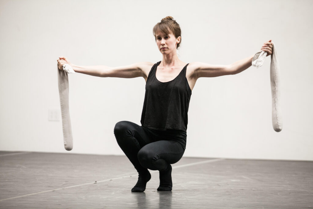 Elise Cowin crouches near to the ground, balancing on the balls of her feet, posture upright. Her arms are extended to the side, holding tubes of fabric weight at the ends. She wears all black; her light brown hair is pulled into a loose bun.