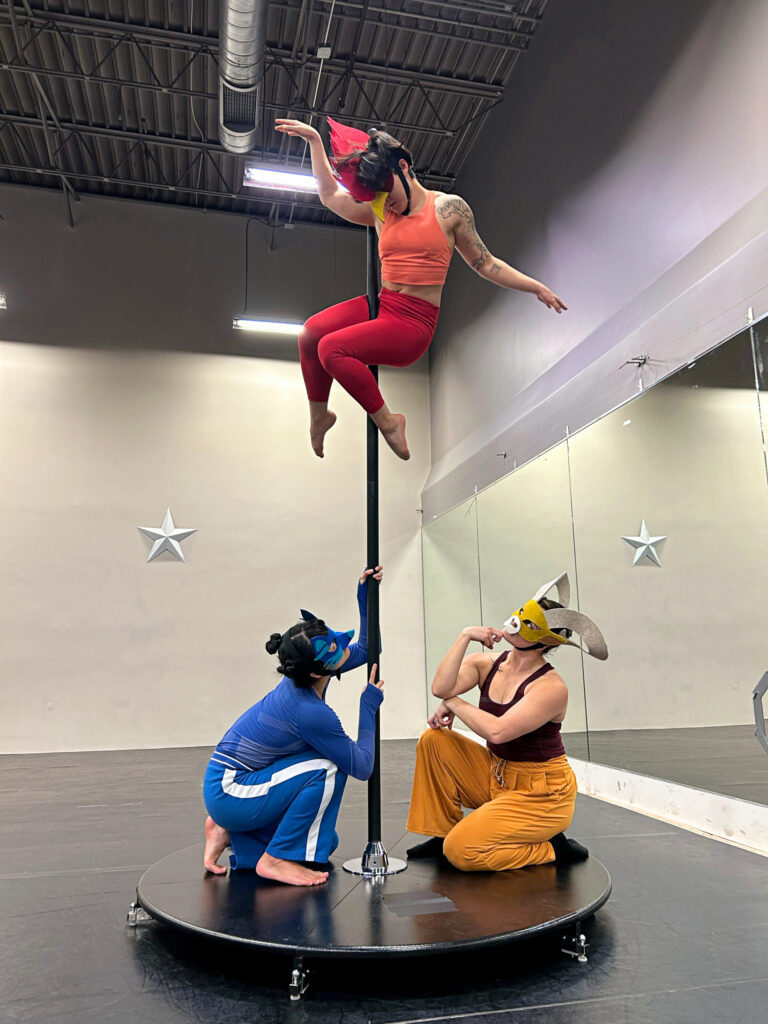 A dancer dressed in orange, wearing a mask evocative of a rooster, balances several feet overhead on a pole. Dancers in blue and yellow, masks evoking a fox and ram, crouch below, looking up at the cockerel.