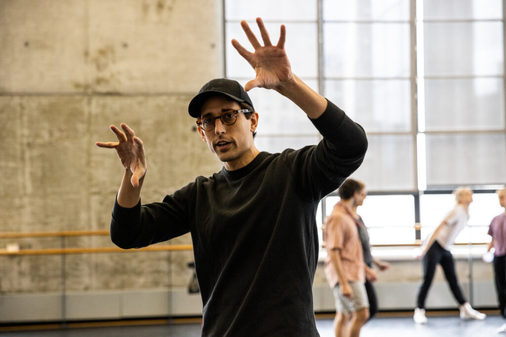 Justin Peck raises both hands above shoulder height, fingers splayed as he illustrates an idea. He wears a long sleeved back shirt and a ball cap. In the background, dancers in rehearsal gear confer with each other.