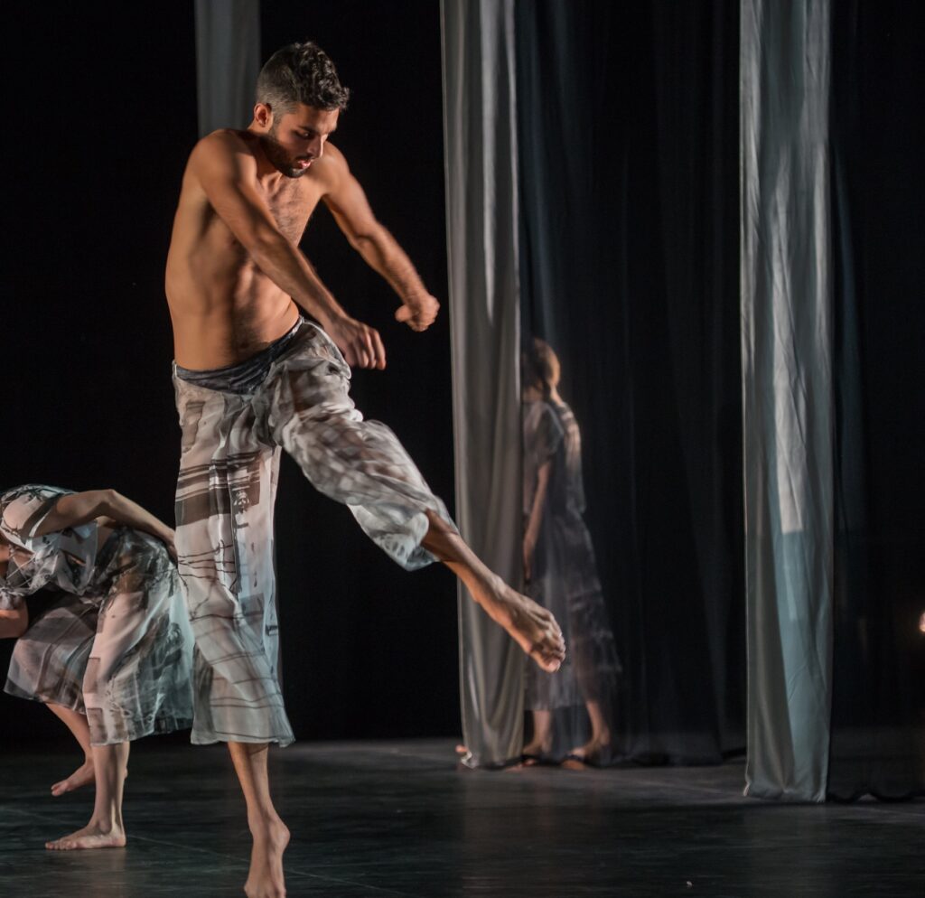 A bare chested Marc Crousillat moves through Trisha Brown's choreography. His torso twists to the left, shoulders rising and hands forming loose fists as his left leg rises in a side parallel attitude. He is barefoot, in a pair of semi-translucent grey pants.