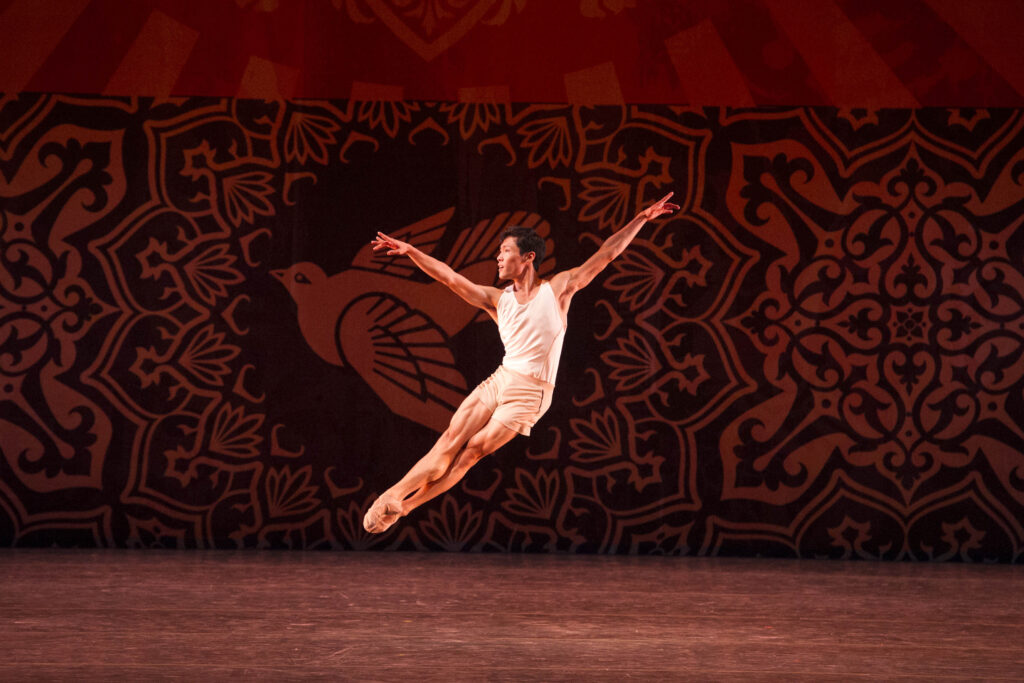 Shimon Ito is caught mid-assemblé, arms floating easily just above his shoulders. He waers lightweight looking shorts and a tank in matching white, and ballet slippers. A warm-toned backdrop illustrating a bird in flight hangs upstage.