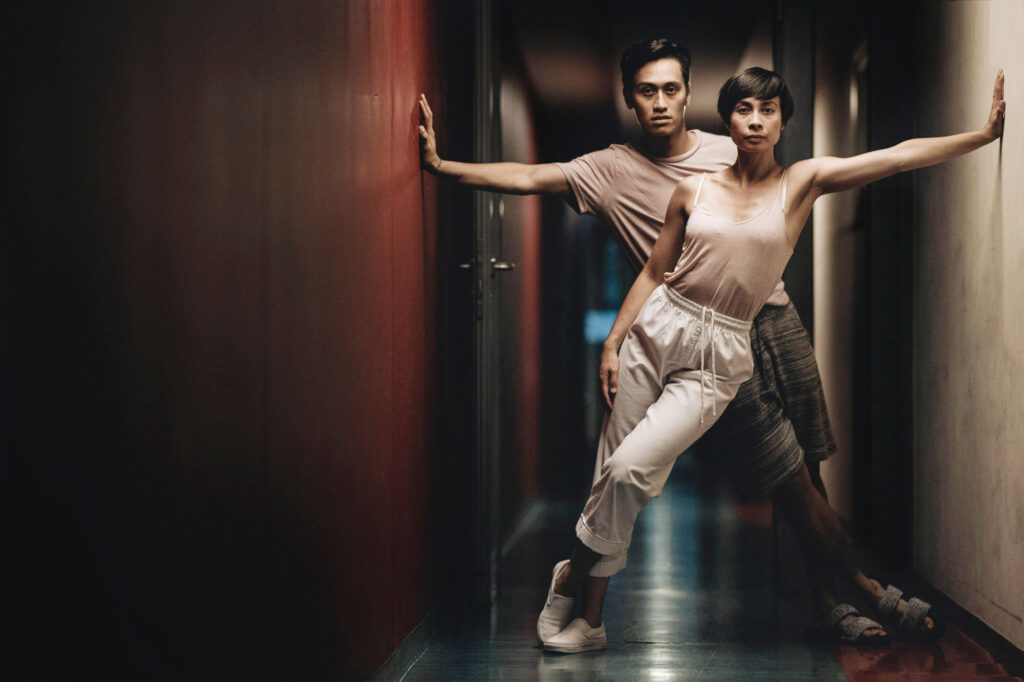 Keone and Mari Madrid face forward as they lean against opposite walls, weight supported on an outstretched arm. Their bodies form an upside-down V intersecting at the center of a narrow hallway. Both gaze solemnly at the camera.