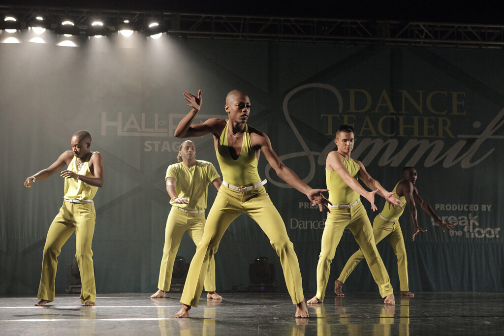 Martha Nichols, a slender Black woman with a shaved head, performs with a group, all wearing the same chartreuse jumpsuits. Her shoulders rise as her hands angularly shape the space, moving through a shallow plié, stance wide.