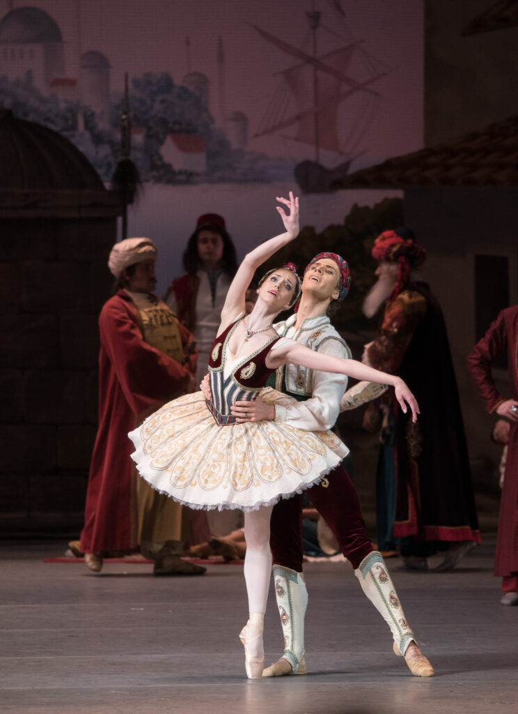 Margarita Shrainer balances in croisé attitude back on pointe, partner standing behind her and supporting her at the waist. Her working side arm is in high fifth, wrist bent so her palm faces outward; the other is elegantly extended side. She wears a pale classical tutu with a darker bodice. Corps members mill in the background.