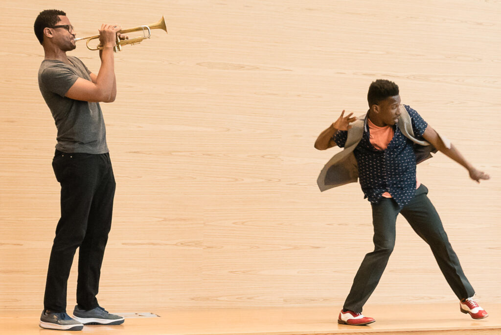 Omari Mizrahi moves through a turned in plié, one leg starting to reach straight along the ground. His open vest flies behind him with his motion as he looks in the direction he's moving. A man playing a trumpet stands behind him.