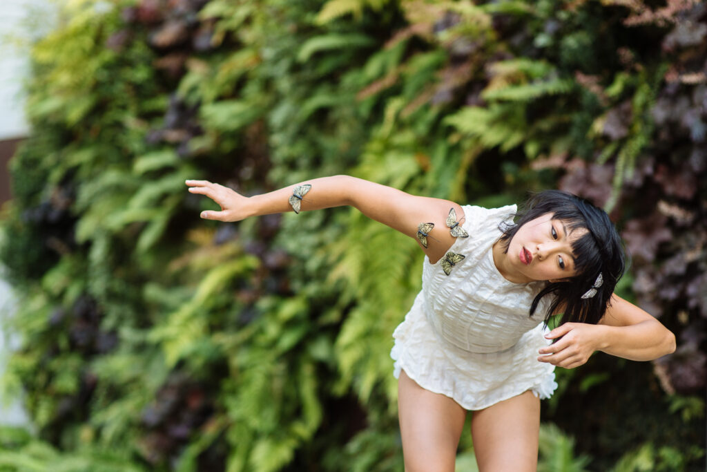 Ching Ching Wong poses against a backdrop of greenery. Butterflies alight on her arm, gently extended to the side, and on her head. She is in a gentle plié, torso tipped forward as her head tips to one side. She wears an airy white romper.