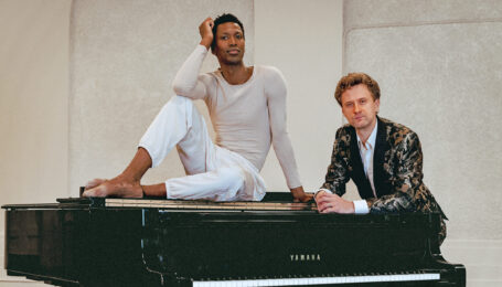 a male dancer sitting on top a grand piano with another man standing behind it
