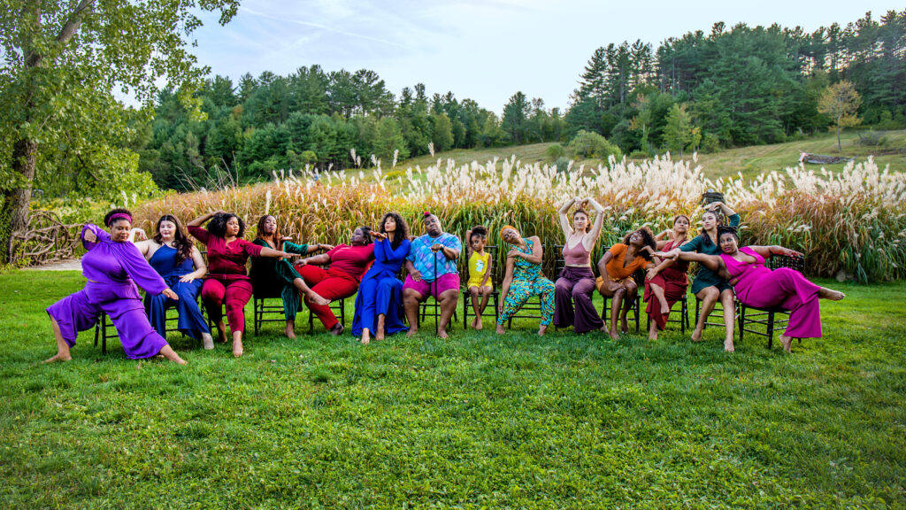 Over a dozen dancers of various ages and body types are seated in a semi-circle, taking various poses from their chairs. The bright jewel tones of their clothing leaps out against the green grasses around and behind them.