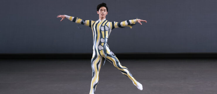 KJ Takahashi is caught mid-air in a temps levé, one leg extended to a 45 degree arabesque, both arms rising at shoulder height. He is alone onstage, wearing a unitard with squiggly patterns.