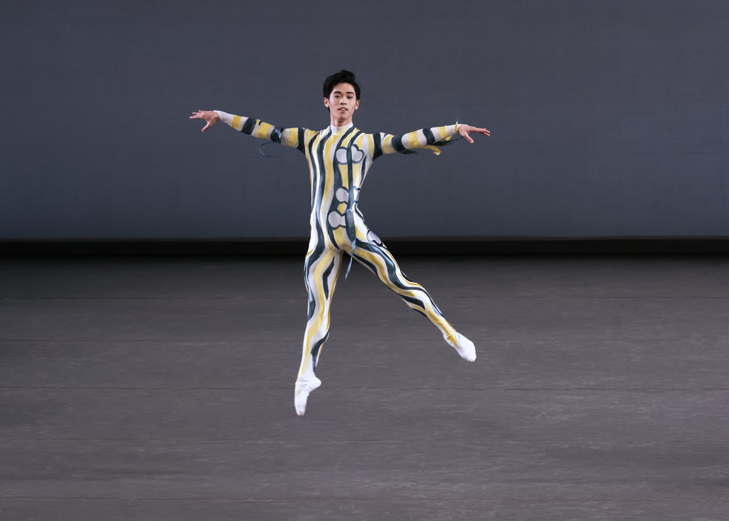 KJ Takahashi is caught mid-air in a temps levé, one leg extended to a 45 degree arabesque, both arms rising at shoulder height. He is alone onstage, wearing a unitard with squiggly patterns.