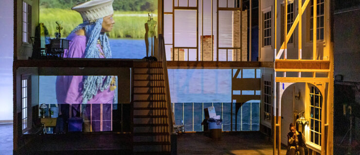 Film is projected over a set that suggests the skeleton of a two story house. A Black woman sits downstage left, under an archway, chin in hand as she stares into the middle distance. The projection shows an older Black woman with cerulean blue braids standing in profile before sky blue water and long green grass.