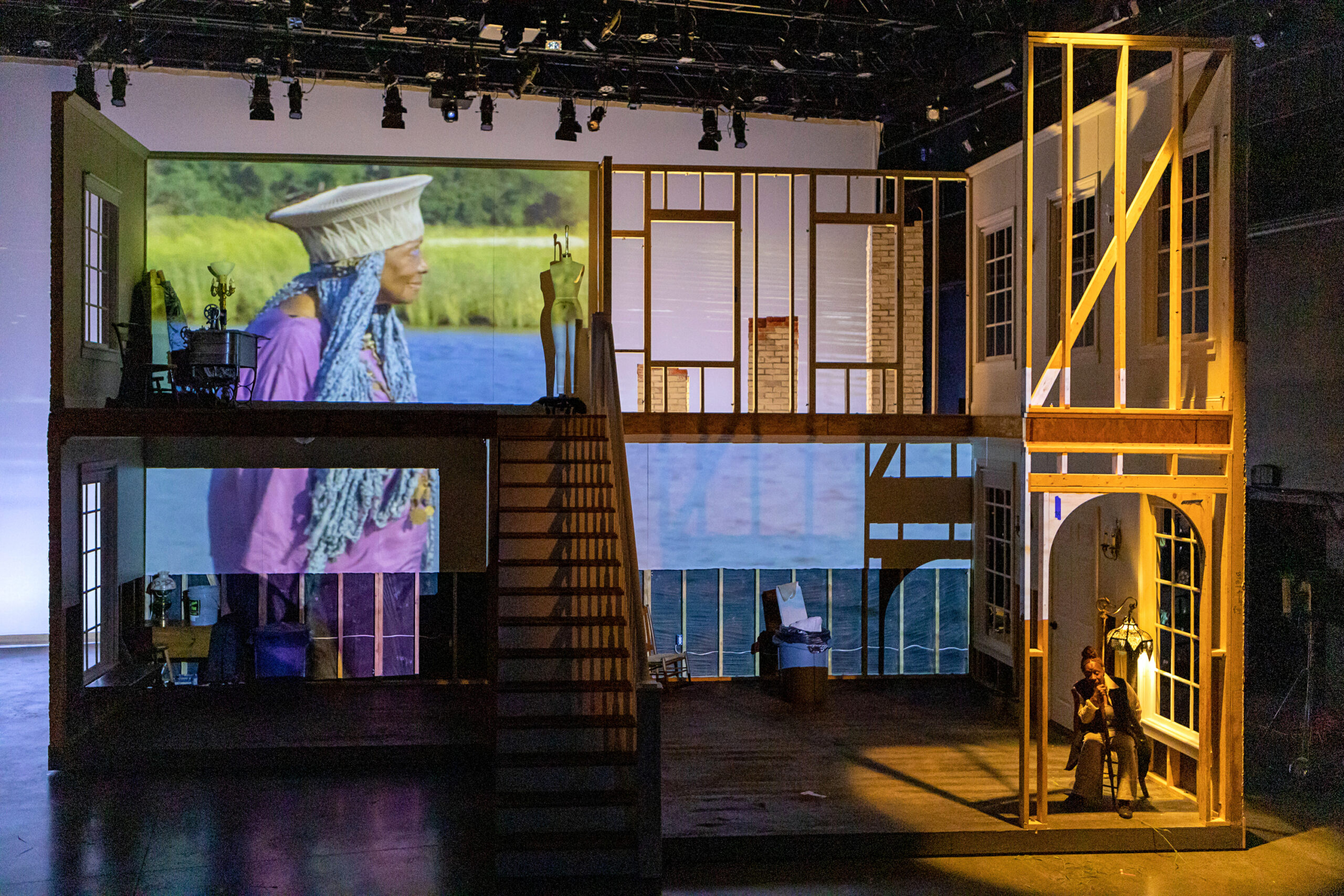 Film is projected over a set that suggests the skeleton of a two story house. A Black woman sits downstage left, under an archway, chin in hand as she stares into the middle distance. The projection shows an older Black woman with cerulean blue braids standing in profile before sky blue water and long green grass.