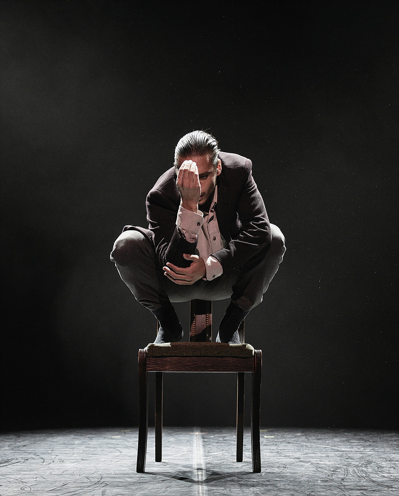 Mario Bermudez Gil crouches atop a dark chair. He wears most of a suit. His right hand covers half of his face; his left hand cups his right elbow, supporting it.