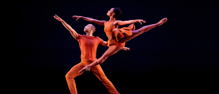 On a dark stage, a female dancer in a warm orange leotard and short skirt leaps beside a lunging male dancer dressed in a short sleeved shirt and fitted pants in the same color. Both look over their front arms, in first arabesque, towards stage right.