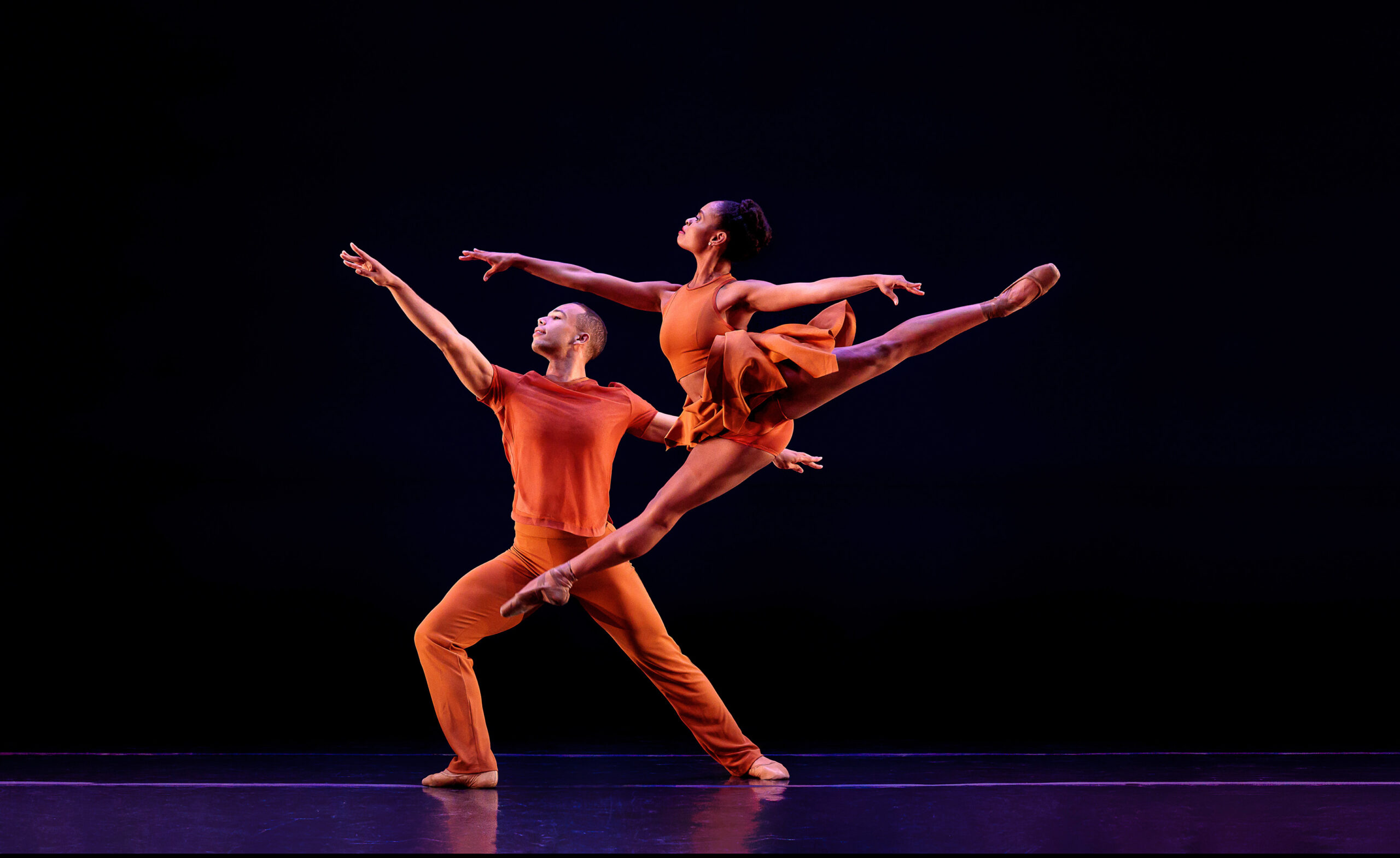 On a dark stage, a female dancer in a warm orange leotard and short skirt leaps beside a lunging male dancer dressed in a short sleeved shirt and fitted pants in the same color. Both look over their front arms, in first arabesque, towards stage right.