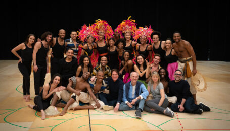 A large group of artists, some wearing flamboyant feathered headpieces, poses on a basketball court.