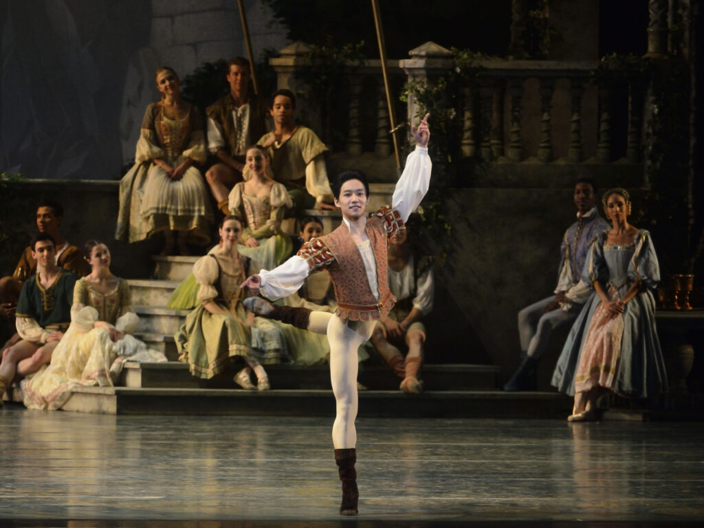 Zhiayo Zhang balances in croisé attitude back, arms in a wide fourth. His manner is one of ease and control. He wears black boots, off-white tights, and a brown and white flowing tunic. In the background, corps members in peasant dresses sit and watch from a set of steps.