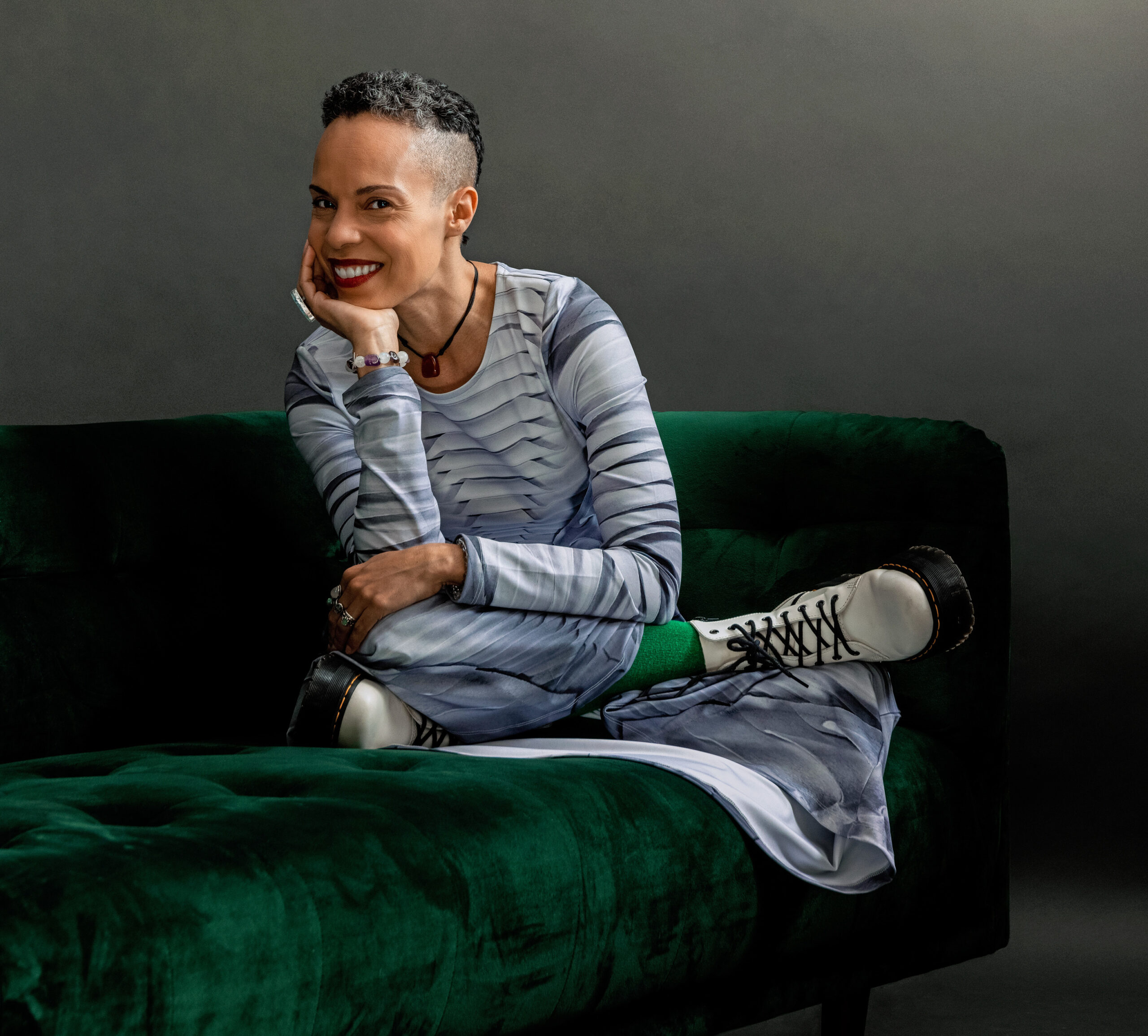 Tai Jimenez grins at the camera as she sits, legs folded under her, on a green velvet couch. She rests her chin in her hand, elbow propped on top of her knee. Her grey and black hair is shaved to a fade on the sides, the tight curls short on top. She wears a grey striped dress, white sneakers with black laces, and bright green socks.