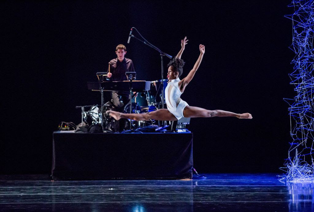 Paige Fraser is caught midair in a serene leap, her arms pushing back behind her torso overhead. She wears a stylized white leotard with stripes of mesh. In the background, a musician is seated at a drumkit atop a raised platform.