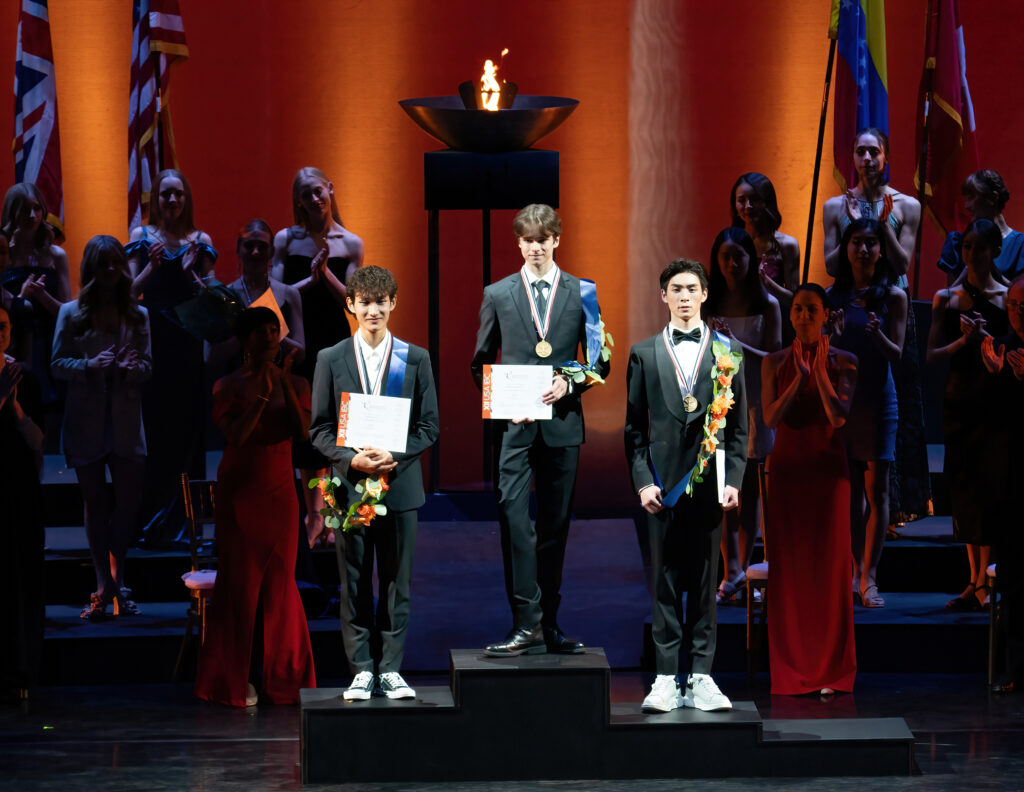 •	Three young men stand on an Olympic-style podium, wearing formal suits and a sash of flowers around their left shoulder. They each wear a medal around their neck.