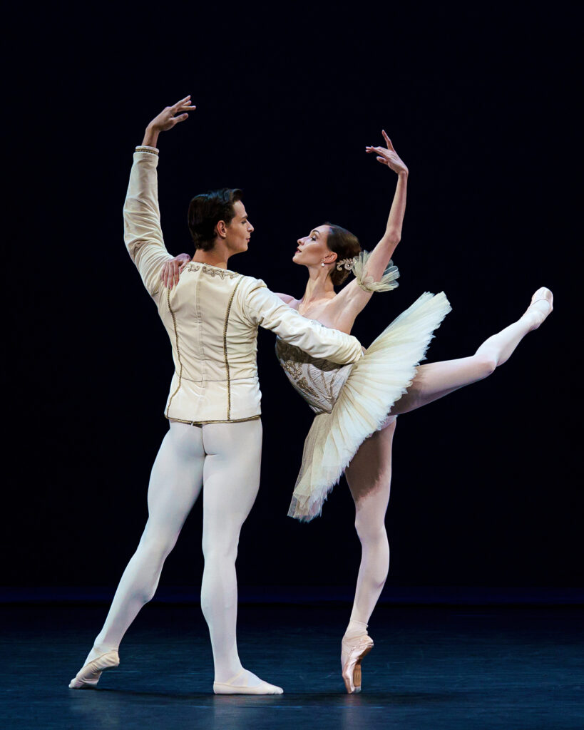 Olga Smirnova balances in attitude back on pointe. She is show in profile, her back arm in high fifth, the other resting on the shoulder of her partner. He faces upstage as he poses in tendu back, supporting her around the waist while his back arm mirrors hers in high fifth.