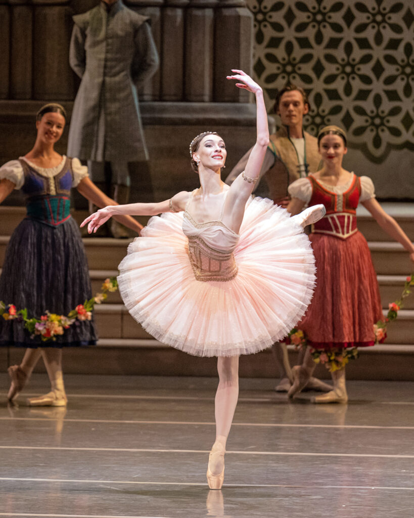 Olga Smirnova balances in back attitude, downstage arm raised in high fifth. She wears a pale pink and gold classical tutu. Corps dancers in long dresses holding flower gardens pose upstage in B-plus.