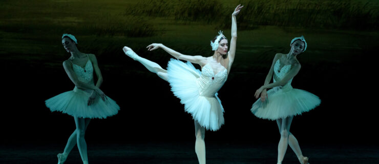 Olga Smirnova as Odette in Swan Lake. She balances in first arabesque on pointe, her front arm raised beside her ear, eyes downcast. She wears a white feathered tutu, pink tights, and matching pointe shoes. Upstage, similarly costumed women pose in B-plus, wrists crossed in front brushing their tutus.