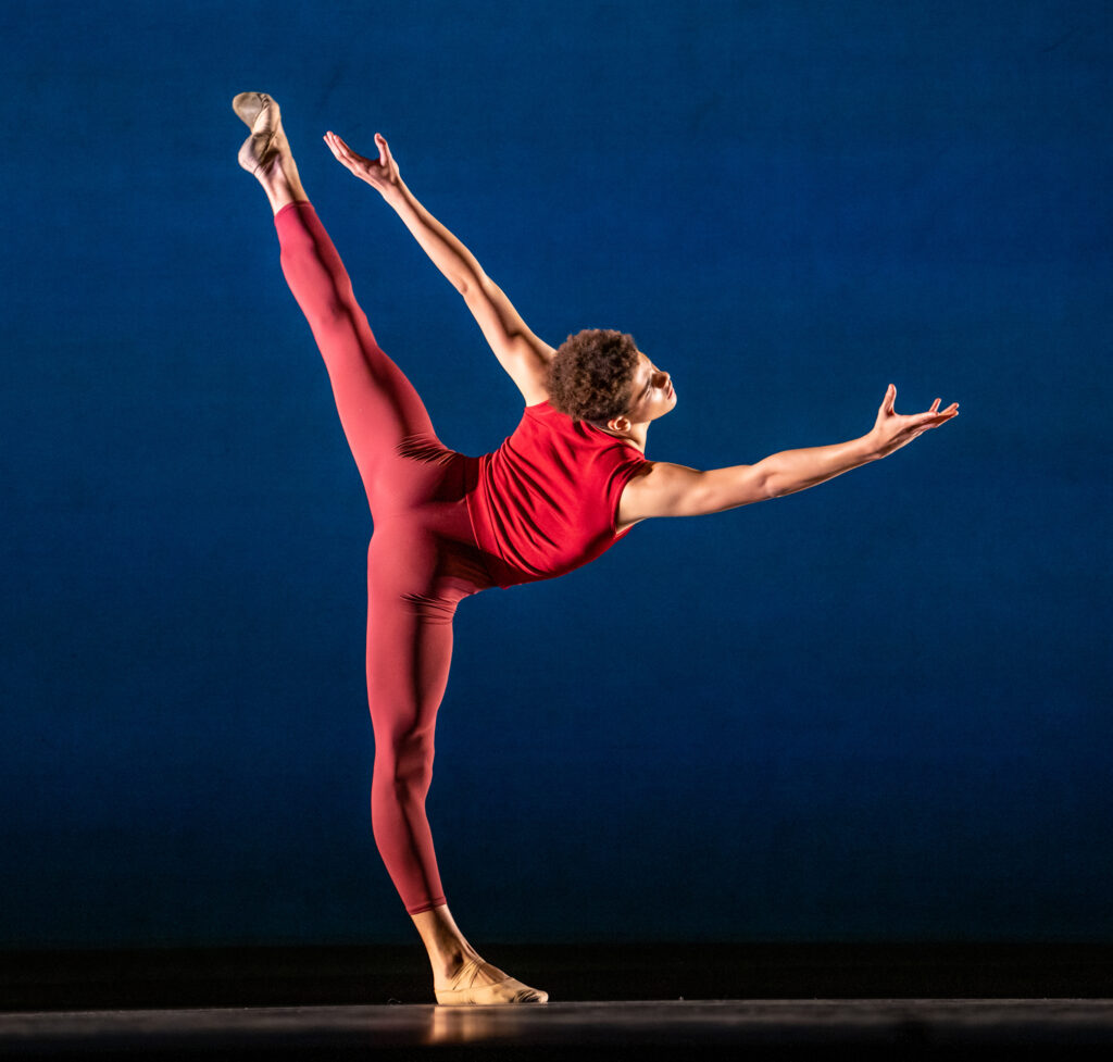 Corbin Holloway wears a red sleeveless top and red footless tights. His body faces the back of the stage and he does a penché with his left leg behind him, reaching his right arm out and stretching his left arm up and back.