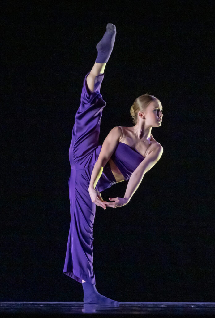 : Isabella Howard does a high grand battement with her right leg and leans her body over to the left, clasping her hands and pulling them downward in front of her body. She wears loose purple pants and a purple tube top and dances in front of a black backdrop.