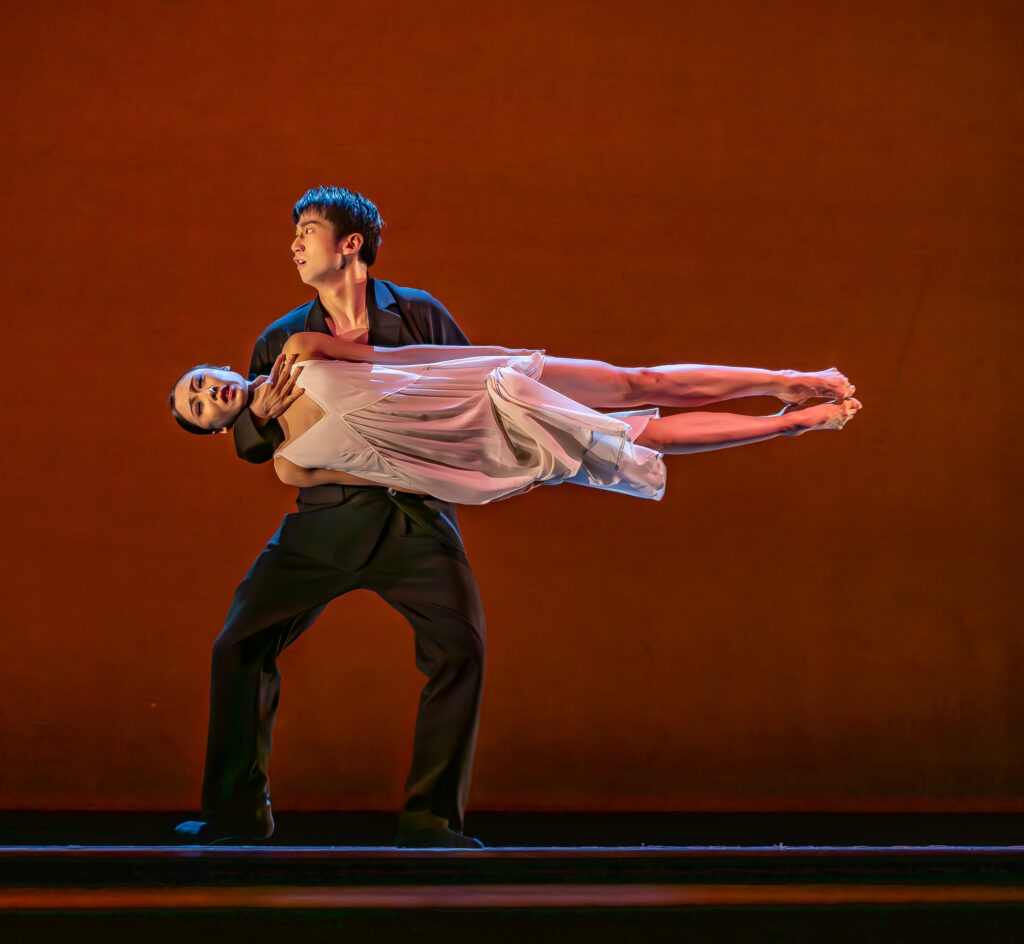 During a performance, Chongzheng Guan holds Xinyue Zhao between her legs and around her neck, propping her horizontally at his waist. She presses her legs tightly together and her arms to her sides. She wears a gauzy white dress, while he wears a black shirt and pants.