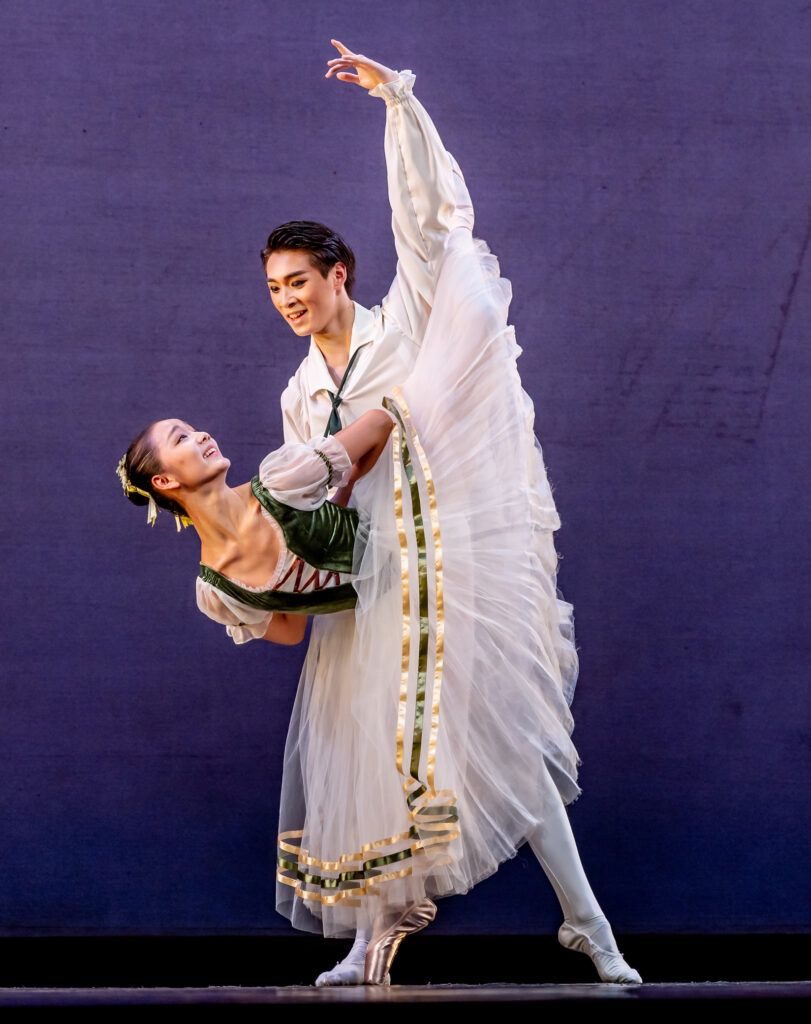 Mengxuan Yan and Zihan Kong perform a classical pas de deux onstage in front of a purple backdrop. She does a penché in attitude with her arms bent and hands clasped behind her back and looks back at Kong, who holds her waist. She wears a white romantic tutu with a green peasant-style bodice, and he wears a white tunic and white tights and ballet slippers.