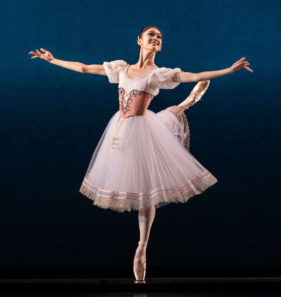 Yunju Lee does a piqué attitude croisé on pointe with her right leg back and her arms open in second with her palms facing up. She wears a light pink peasant costume with a knee-length tutu.