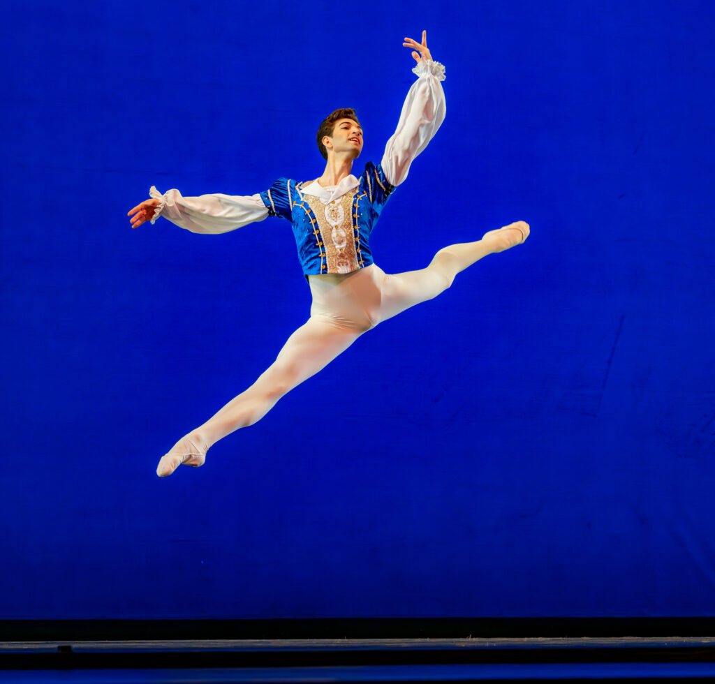 Yuval Cohen performs a sissonne fermé en avant, splitting his legs and holding his arms sin third position. He wears a blue tunic with white billowing sleeves, white tights and white ballet slippers and dances in front of a bright blue backdrop.