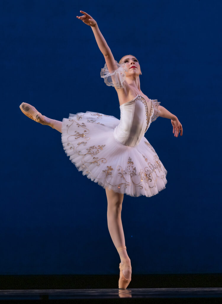 Julie Joyner performs a piqué attitude in croisé during a performance with her left lag in back. She raises her right arm and looks up toward it. She wears a white classical tutu, pink tights and pink pointe shoes and dances in front of a blue backdrop.
