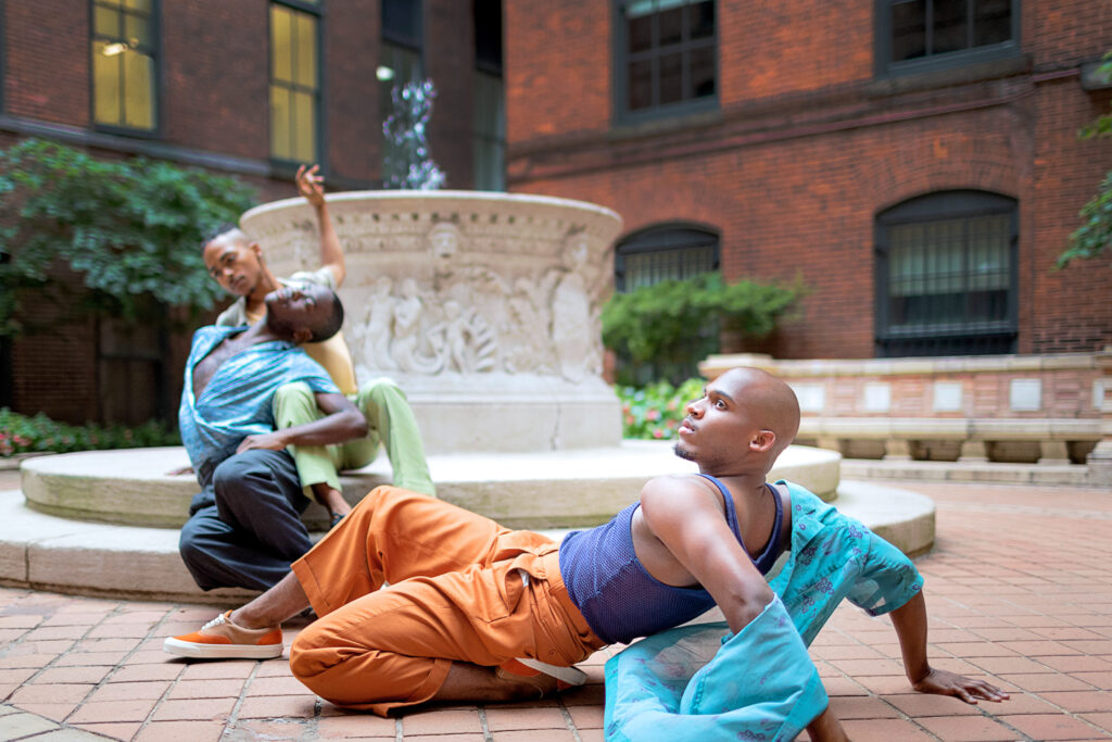 Three Black dancers in pastels and bright colors in an outdoor courtyard. One leans back on their palms, a knee tucked beneath them. The other two sit on the lip of a small fountain, leaning into each other.