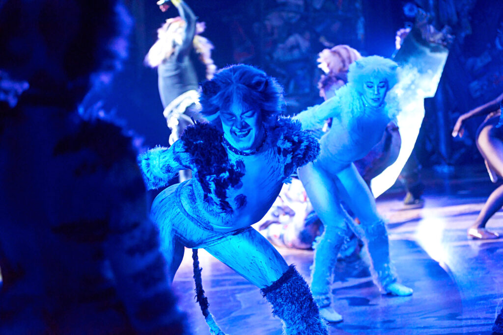 Kolton Krouse performing in full cat costume, hair, and makeup, as seen from the wings. They lean forward, stance wide, arms extended behind them. Other dancers are visible doing the same in the foreground and the background.