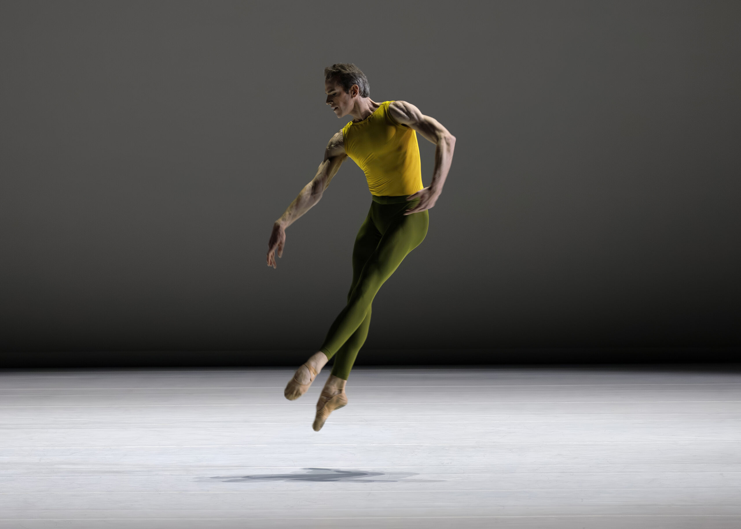 Russell Janzen hovers above the floor, feet crossed in a low fifth. He arches forward slightly, looking out over an arm extended in parallel to his legs. He wears a fitted yellow top and dark green tights. The backdrop is dark grey; he casts a shadow on the pale grey marley.