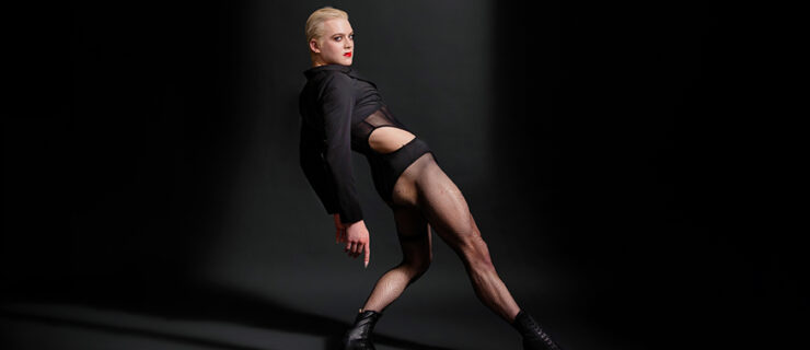 Kolton Krouse poses in profile against a dark backdrop. One leg extends long along the ground, their supporting knee bent, arms draping behind their hips. Their hips thrust forward to create a hinge. Krouse looks over their shoulder to smolder at the camera, short blond hair slicked back and lips painted red. They wear heeled lace-up jazz boots, fishnet tights, and a black fashion leotard with geometric cutouts at the hips and billowy long sleeves.