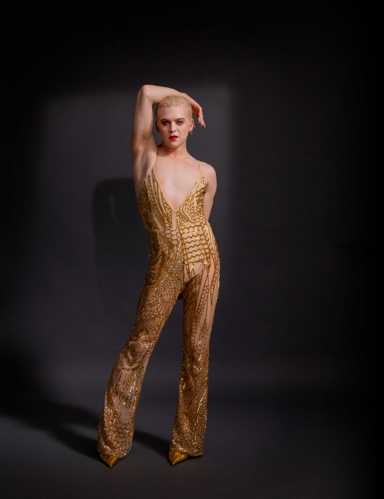 Kolton Krouse poses against a dark backdrop. They sit into one hip, a forearm draped over their head as they look at the camera head on. They wear a lowcut golden jumpsuit. Their short blond hair is slicked back and their lips painted red.