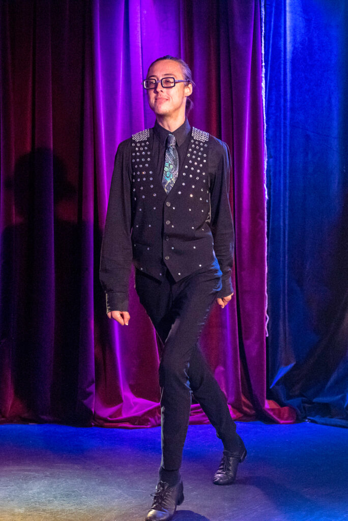 a dancer wearing all black dancing in front of a purple curtain 