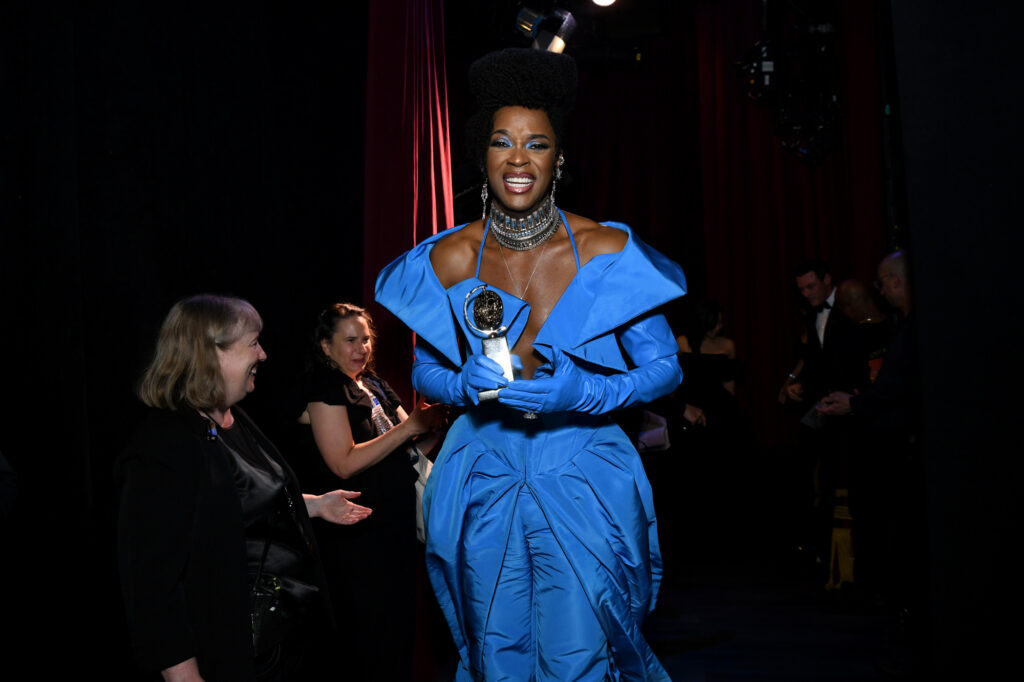 Ghee, a chocolate-skinned Black nonbinary person in.a vibrant blue off-the-shoulder ensemble and matching long gloves, beams as they stand in the wings of a theater, holding their Tony Award.