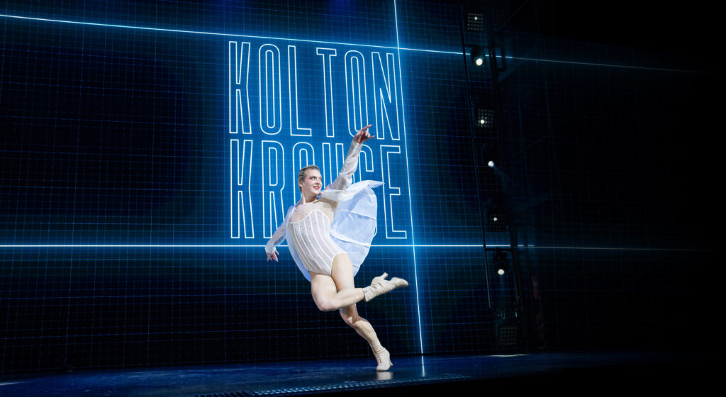 Kolton Krouse flicks a pointed foot over a bent supporting knee, face turned out towards their upraised arm. A black backdrop is illuminated with massive blue letters spelling out "Kolton Krouse." They wear a ribbed white leotard and an unbuttoned long sleeved shirt.