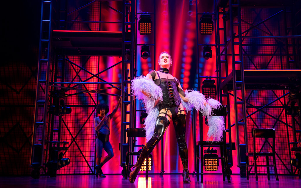 Kolton Krouse takes a wide stance center stage, one shoulder tipped forward and chin raised confidently. They wear a pale feather boa like a shrug, over a strappy black leotard and corset and thigh-high black boots. Scaffolding and lights are visible upstage. The projected backdrop is a mix of pinks, reds, and blues.