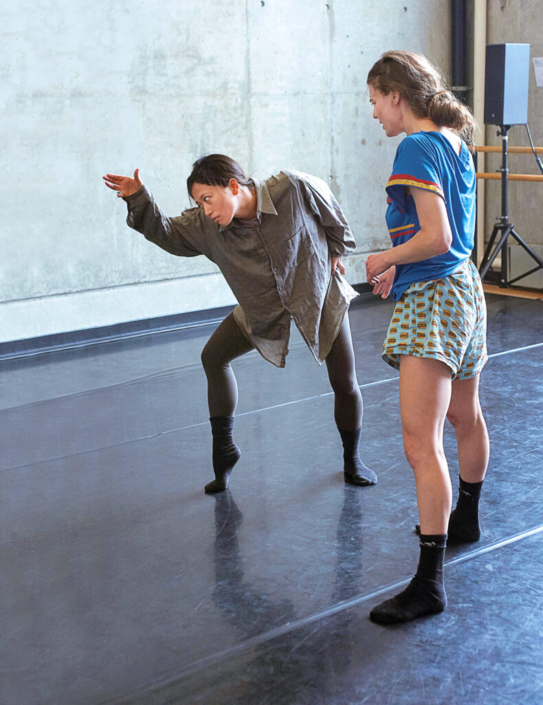 Two dancers work in a studio in comfortable rehearsal gear. One looks on as the other leans forward, staring intently at at their hand as they extend two fingers. They lean over a foot perched on forced arch.