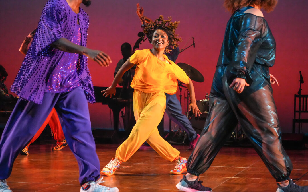 A dancer costumed in bright yellow steps into a wide stance, legs crossed. Her arms are raised at the elbows, hair flying up as she begins to turn. In the foreground, a couple of dancers stride around her. Upstage are musicians.