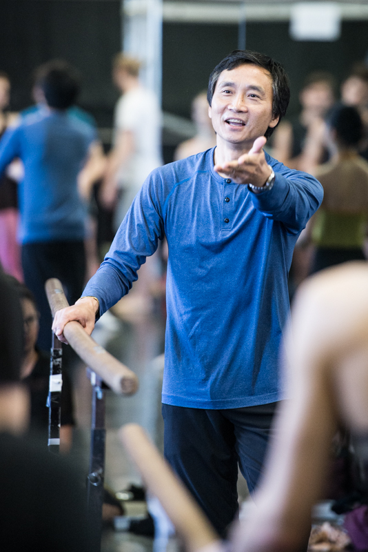 Li Cunxin stands with one hand on the barre. He reaches his outside arm forward, palm up.