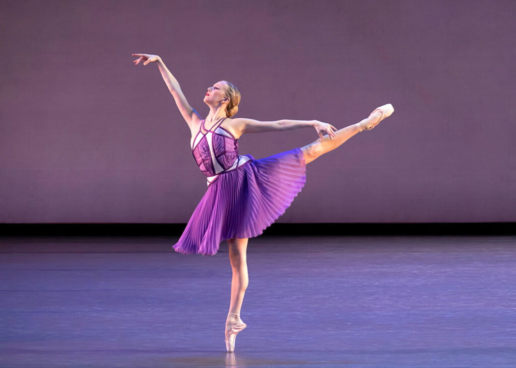 a female dancer wearing a knee-length purple dress performing first arabesque on stage 